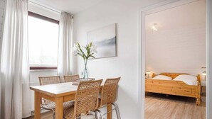 A room with a wooden dining table and chairs, a bed with a wooden frame, and a painting on the wall. - Helle Wohnung mit 2 Schlafzimmern (Sankt Peter-Ording)