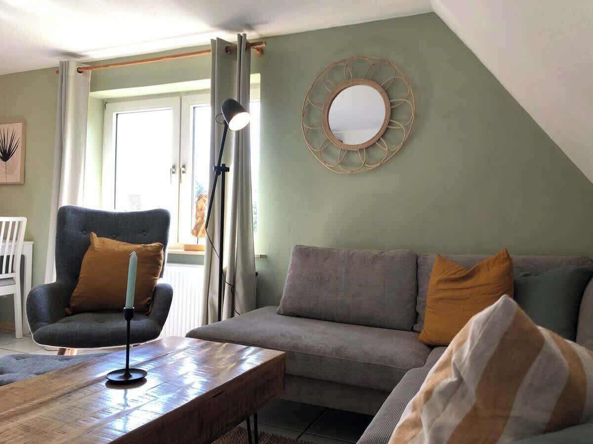A living room with a sofa, armchair, and a coffee table. There is a lamp, a mirror on the wall, and a door with curtains.