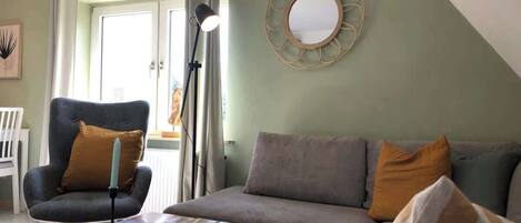 A living room with a sofa, armchair, and a coffee table. There is a lamp, a mirror on the wall, and a door with curtains.