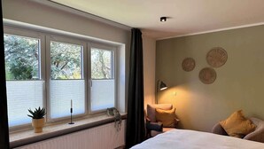 Un dormitorio con un ventanal grande, una cama, una silla y una mesita de noche.