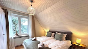 A bedroom with a sloped ceiling, a bed with a grey blanket and pillows, a bedside lamp, and a window with a view of trees. - Quiet, comfortable apartment near the beach (Sankt Peter-Ording)
