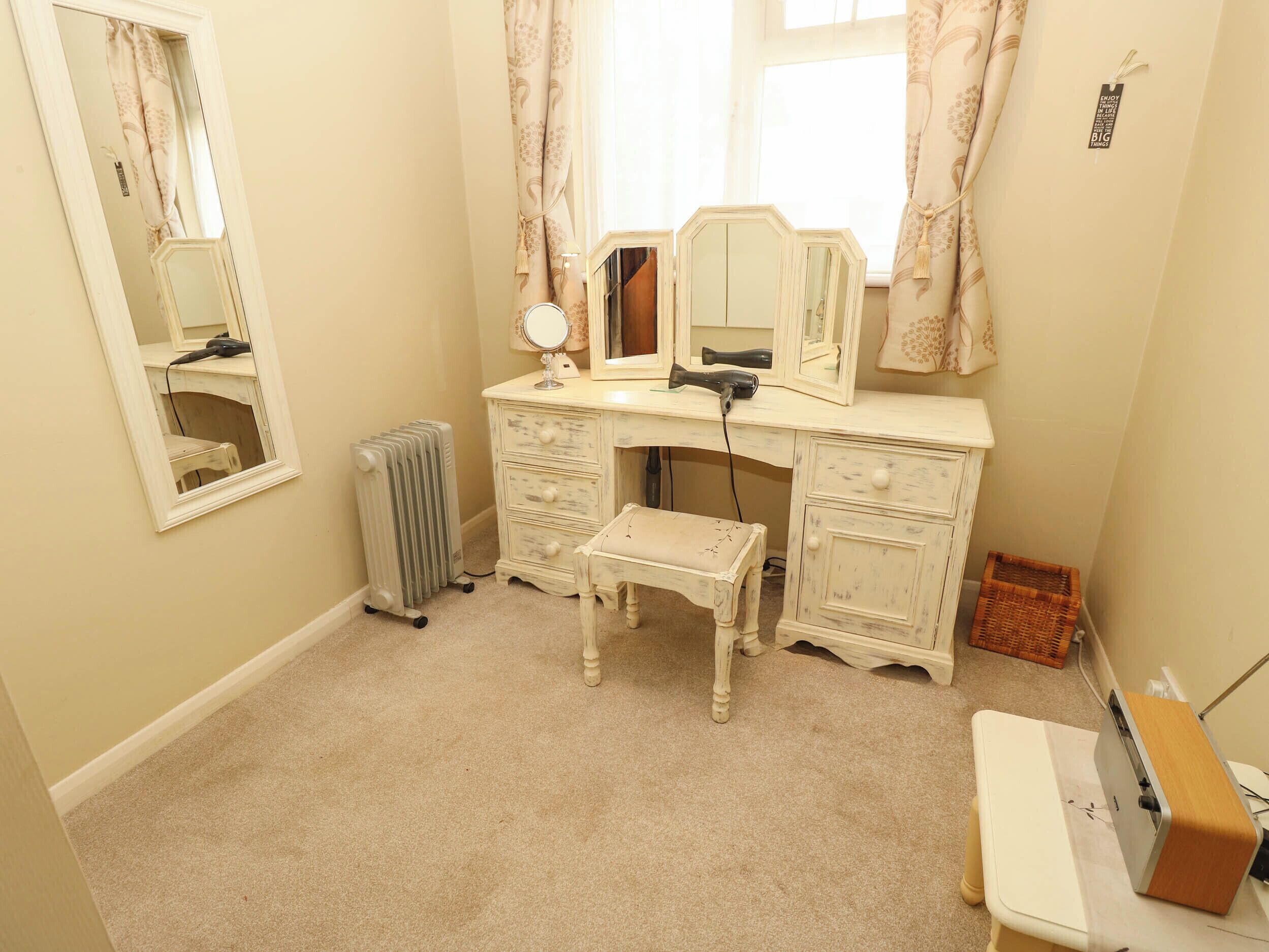 A small, well-lit room with a vanity area, including a mirror, a hairdryer, and a radiator. There is a bench and a small table with a sewing machine.