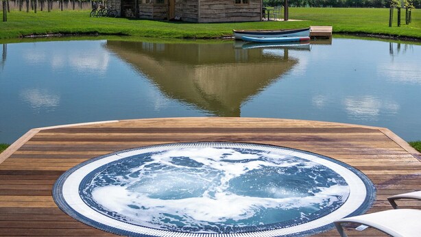 A thatched-roof house with a hot tub on a wooden deck, a small boat, and a calm pond. - Wilderness Reserve - Hayjack (Saxmundham)