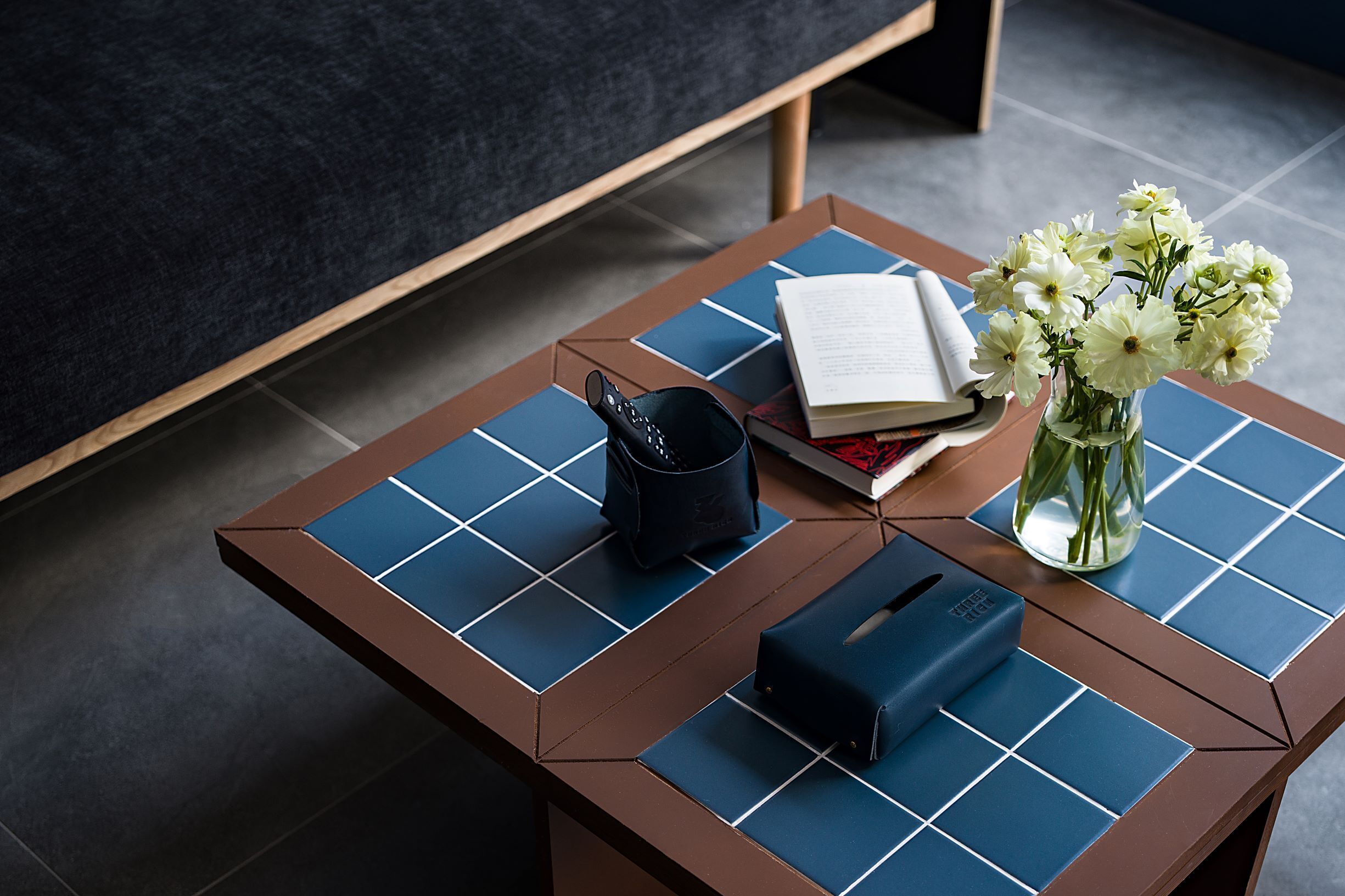Una habitación moderna con una mesa de centro marrón con un estampado azul de cuadrícula, un control remoto negro, un libro y un jarrón con flores blancas.