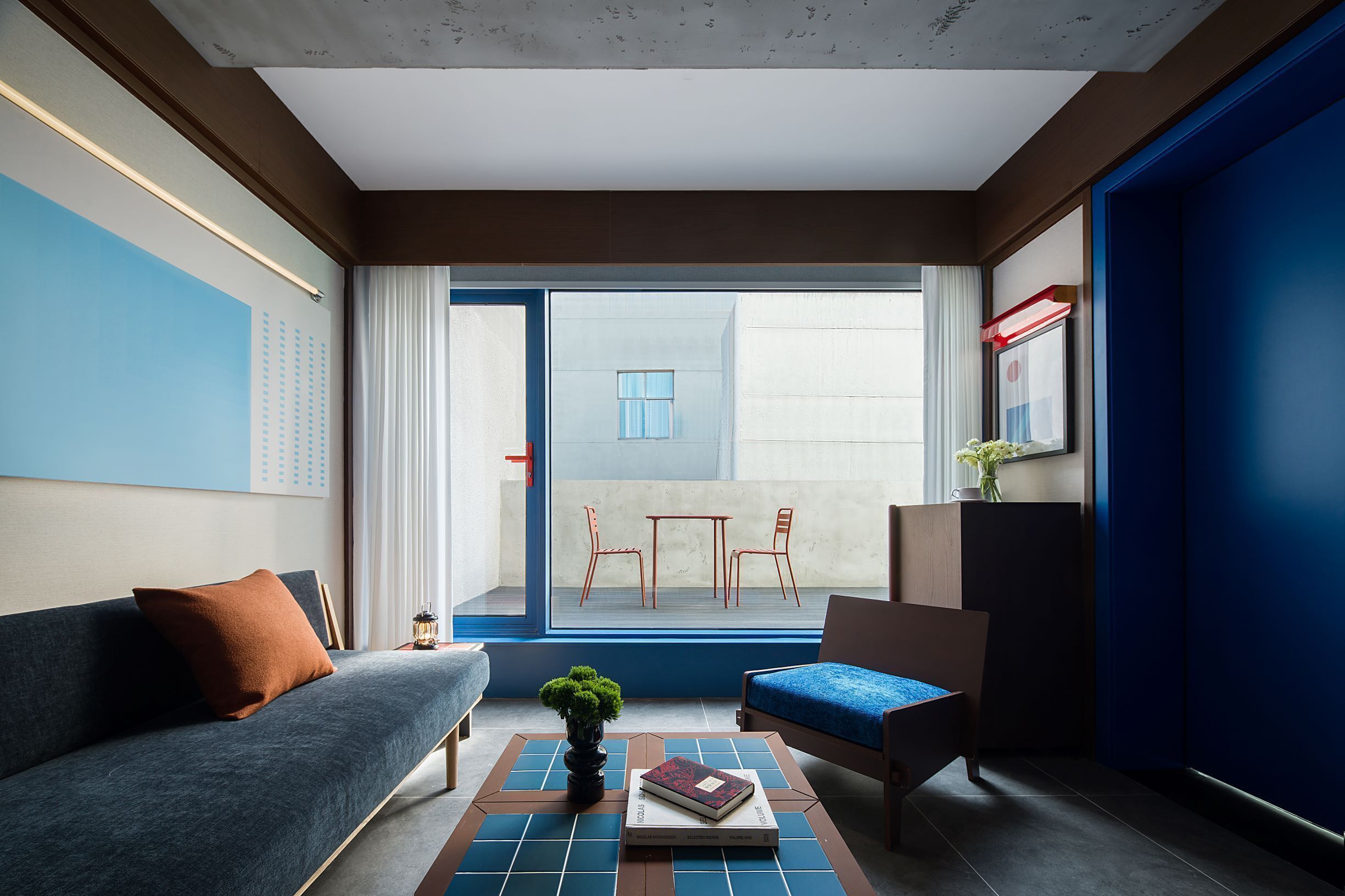 A modern hotel room with a sofa, a small table with a plant and a book, a chair, and a view of the outside balcony.
