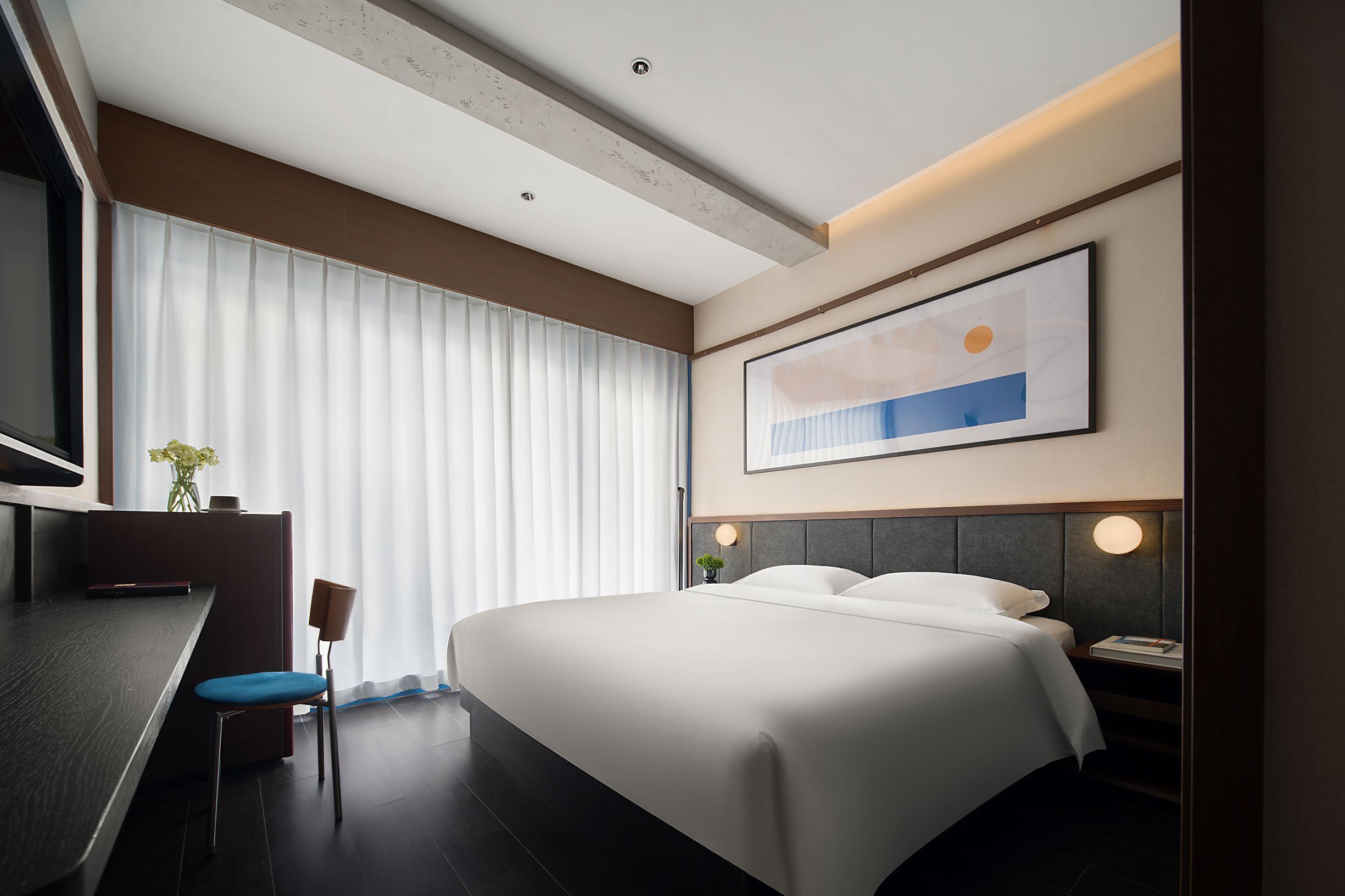 A modern hotel room with a large bed, a desk, and a chair. There is a large window with blinds, a painting on the wall, and a small plant on the desk.