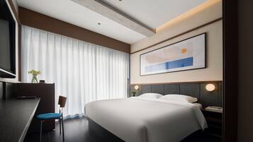Habitación de hotel moderna con una cama grande, un escritorio y una silla. Cuenta con un ventanal con persianas, un cuadro en la pared y una planta pequeña en el escritorio.