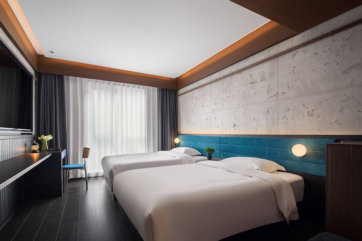 A hotel room with two beds, a desk, a chair, and a large window with curtains.