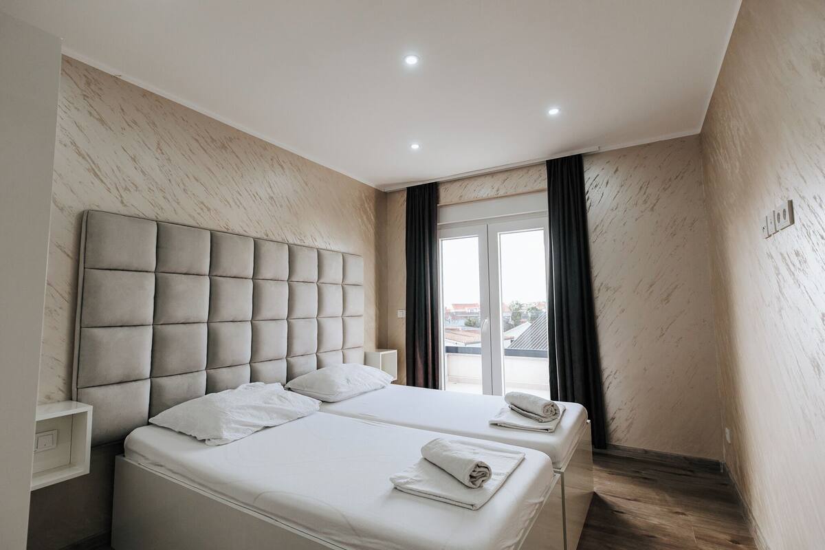A modern hotel room with a large bed, a headboard, a bedside table, and a window with curtains.
