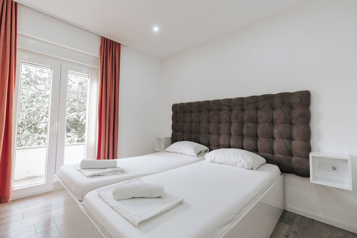 A hotel room with two beds, a large window with curtains, a tufted headboard, and a bedside table with a lamp.