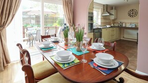 A dining area with a wooden table set for four, surrounded by chairs. A kitchen is visible in the background with a clock on the wall. - White Lion Retreat (Cromer)