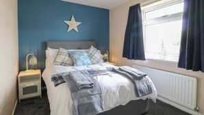 A neatly made bed with blue and white bedding, a star decoration on the wall, a nightstand with a lamp, and a window with curtains. - White Lion Retreat (Cromer)