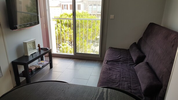 Ett vardagsrum med ett mörkt, runt bord, en lila soffa, en liten svart tv på väggen och en balkong med utsikt över byggnader och träd.