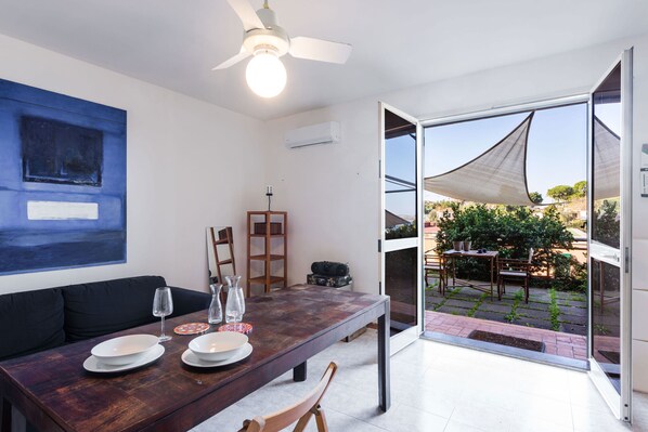 Un coin repas avec une table en bois, des chaises et un ventilateur de plafond. On peut admirer la vue sur l’extérieur à travers une porte vitrée.