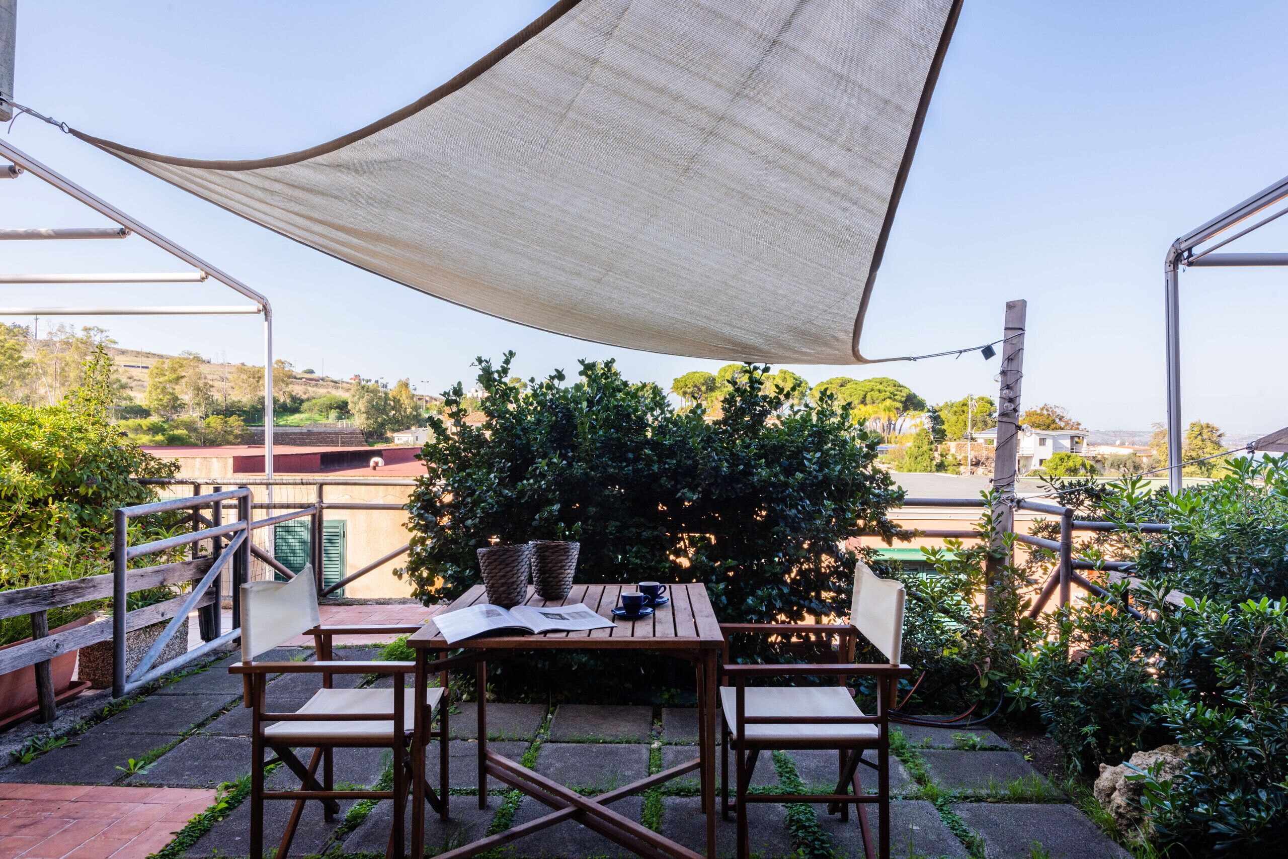 A patio with a table and chairs, a large shade sail, and a view of the surrounding area.