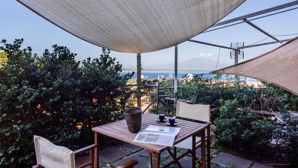 Une table en bois, un journal, un panier en osier et une tasse placés sous un grand auvent, avec vue sur un jardin et la mer.