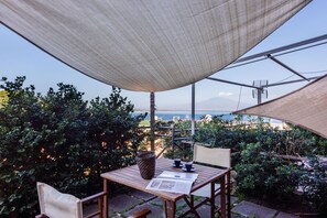 Une table en bois, un journal, un panier en osier et une tasse placés sous un grand auvent, avec vue sur un jardin et la mer.