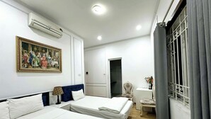 Een hotelkamer met twee bedden, een schilderij, een airconditioner, een bureau met een lamp, een stoel en een raam met gordijnen.