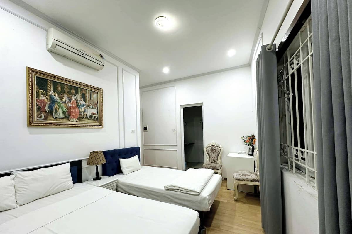 Habitación de hotel con dos camas, un cuadro, aire acondicionado, un escritorio con lámpara, una silla y una ventana con cortinas.