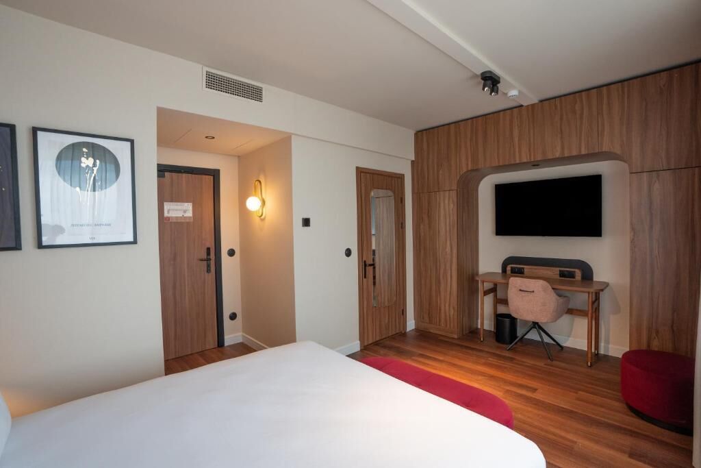 Ein modernes Hotelzimmer mit einem großen Bett, einem Schreibtisch mit Stuhl, einem an der Wand befestigten Fernseher und gerahmten Kunstwerken.