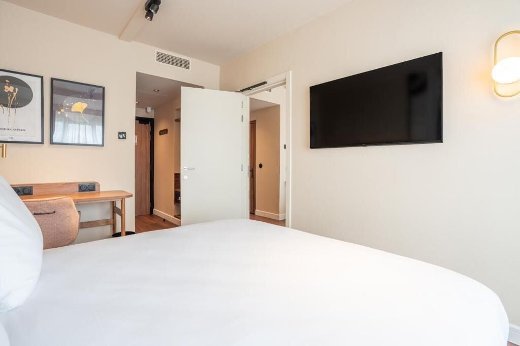 Habitación de hotel con cama, escritorio, silla, televisor montado en la pared y cuadros decorativos.