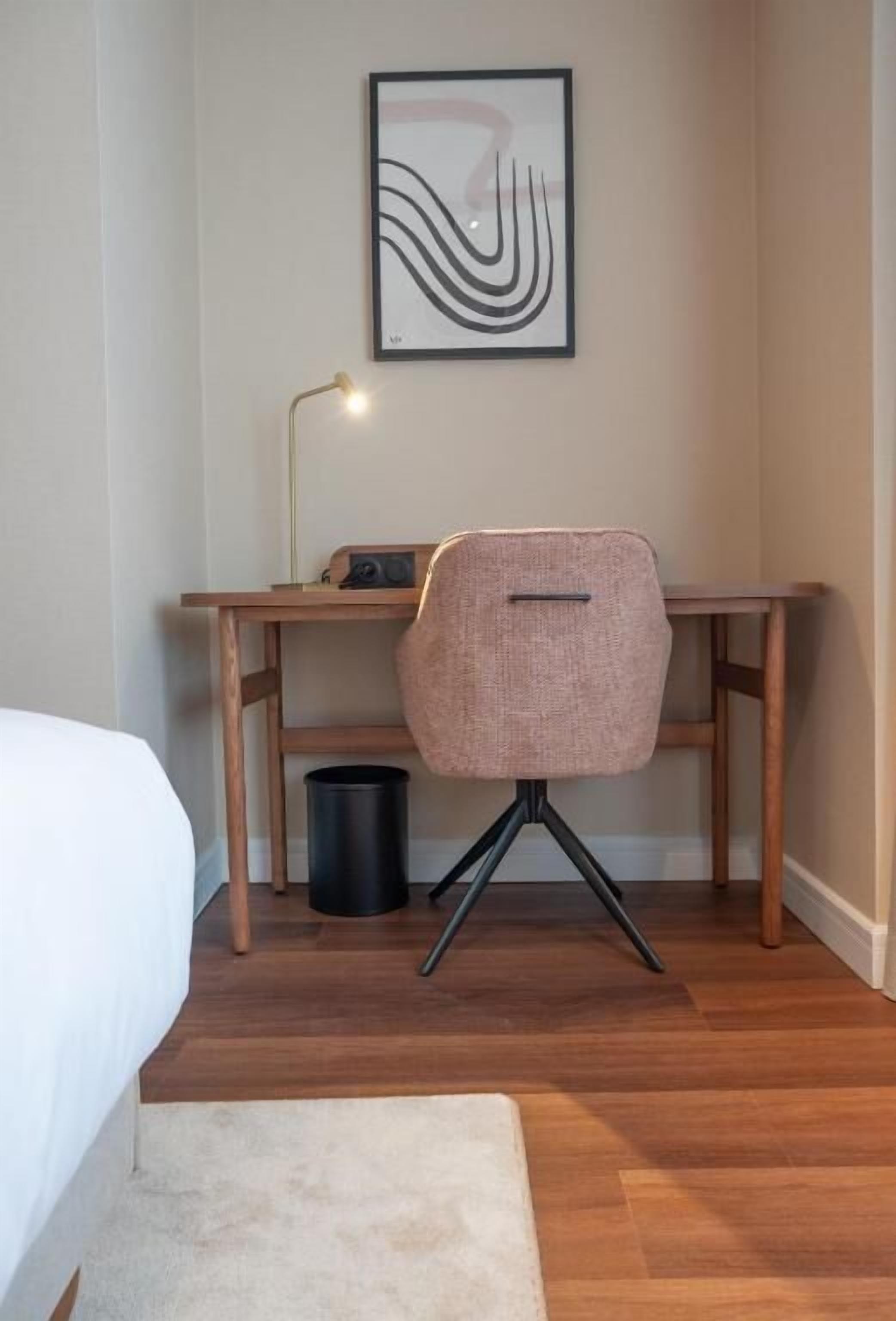 A bedroom with a wooden desk, a chair, a lamp, and a framed artwork on the wall.