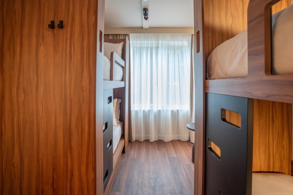 A compact, well-lit interior with a wooden floor, a bunk bed, and a window with sheer curtains.