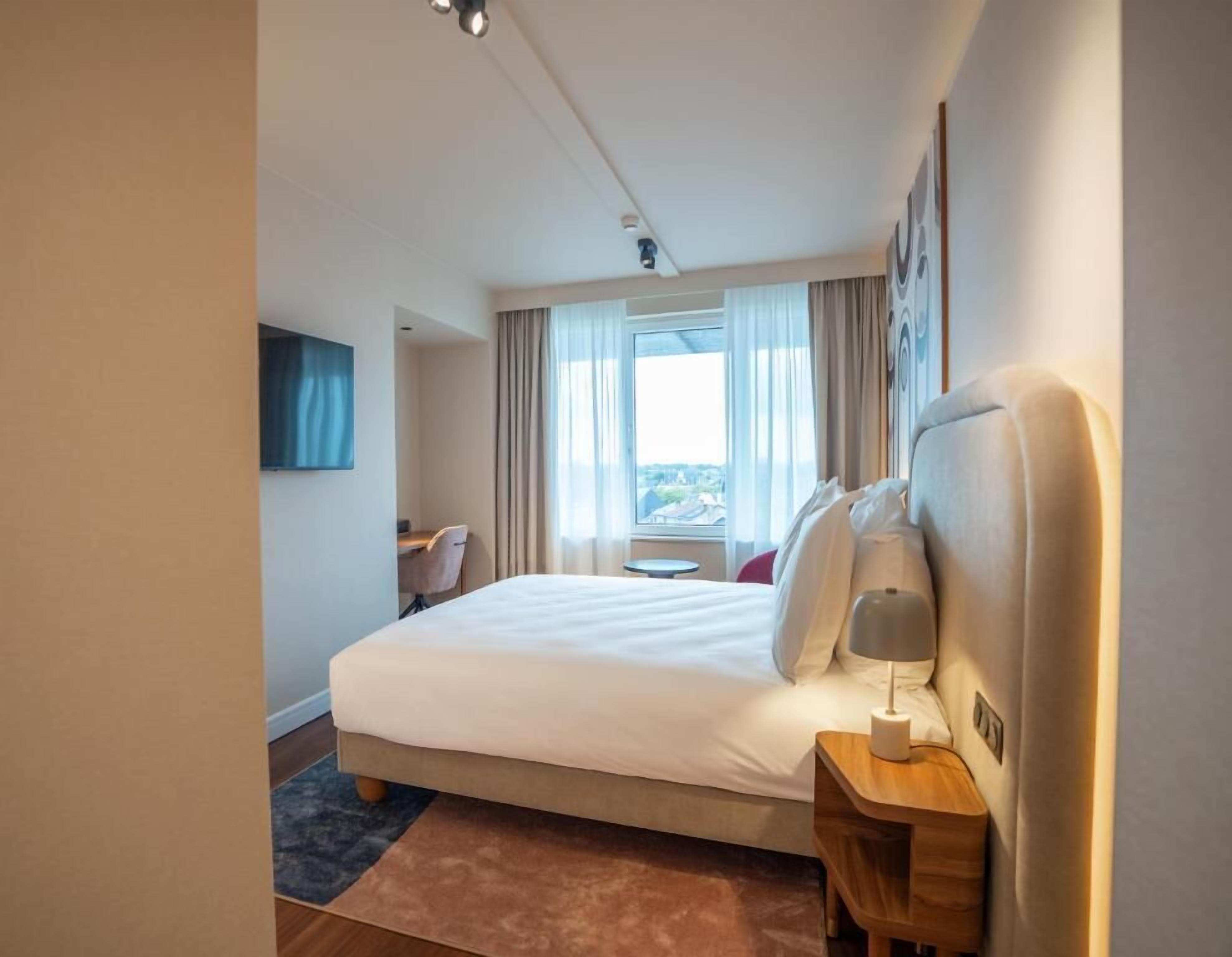 A hotel room with a large bed, a desk, and a view of the city through the window.