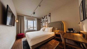 Camera d'albergo con un letto grande, una scrivania in legno, un poggiapiedi rosso e una TV a parete.
