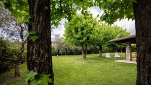 A grassy outdoor area with a stone gazebo, two white lounge chairs, and trees. - Villa Golf in Polpenazze del Garda (Polpenazze del Garda)