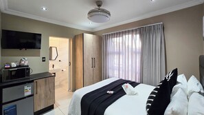 A hotel room with a bed, a TV, a minibar, a bathroom, and a large window with curtains. - Sunset Events & Accommodation (East London)