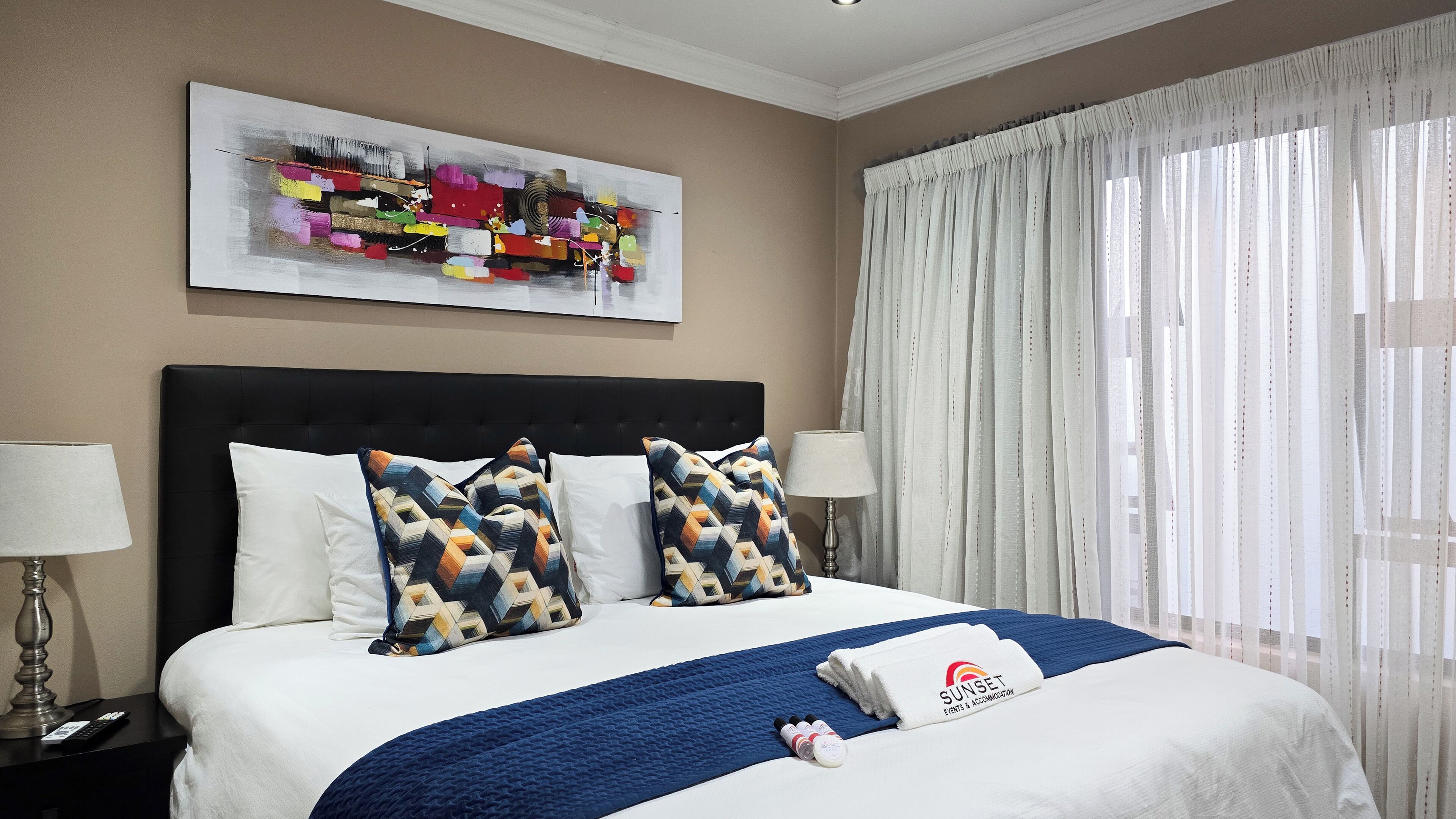 A hotel room with a large bed, two pillows, a blue blanket, a white towel, and a painting on the wall.