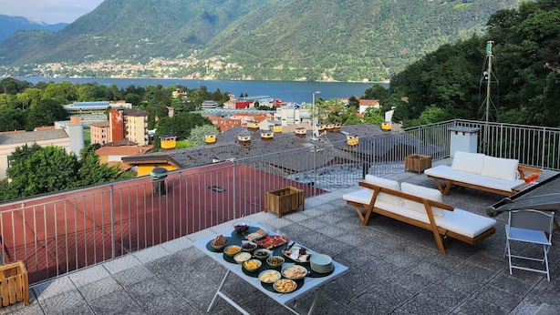 A rooftop terrace with a table set for a meal, overlooking a mountainous landscape and a body of water. - Sky Attic Lake Como (Como)