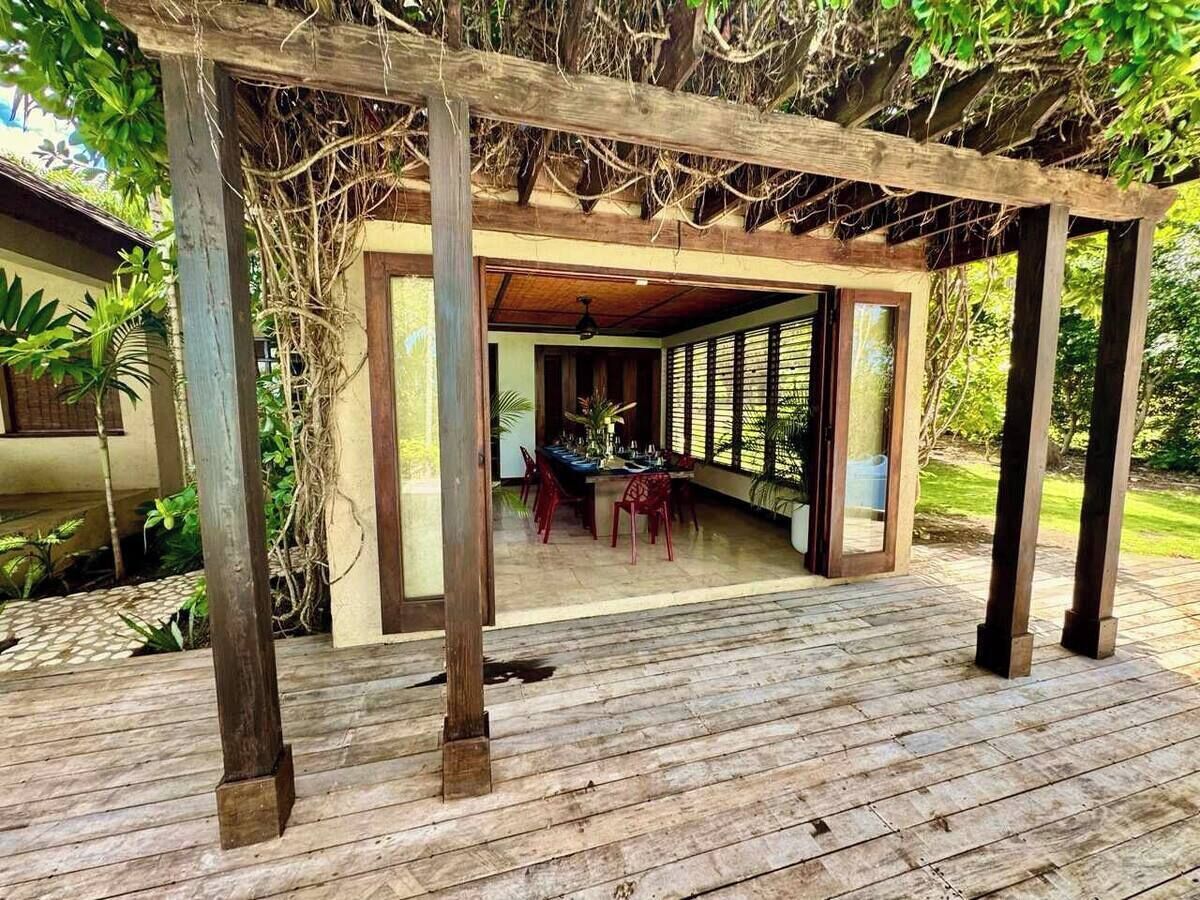 Una terraza de madera con una zona de estar cubierta, una mesa de comedor con sillas y una vista de una casa al fondo.
