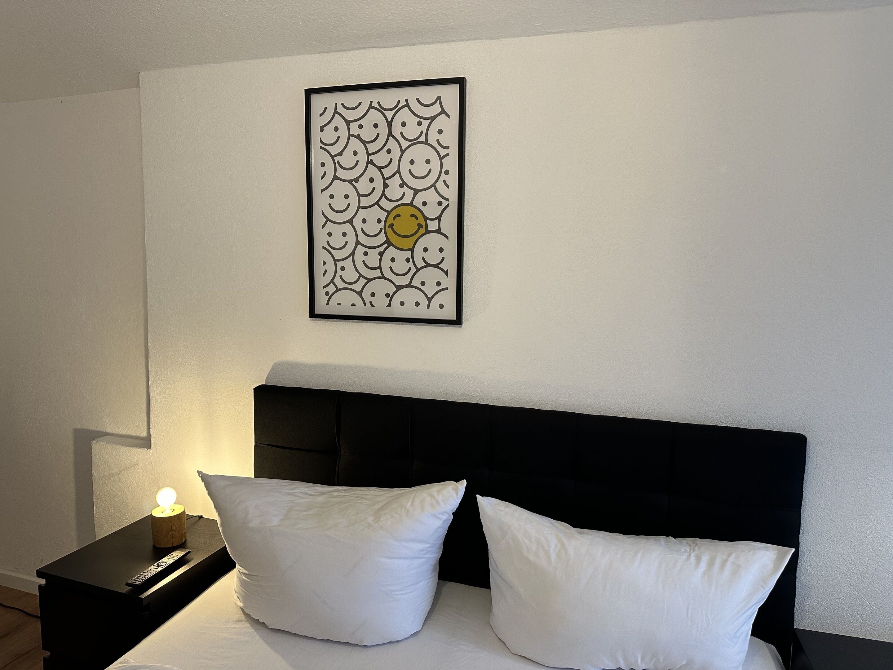 Un dormitorio con cabecera negra, dos almohadas blancas, una mesita de noche con control remoto y un cuadro en la pared con caritas sonrientes.