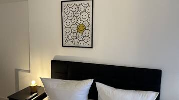 Un dormitorio con cabecera negra, dos almohadas blancas, una mesita de noche con control remoto y un cuadro en la pared con caritas sonrientes.