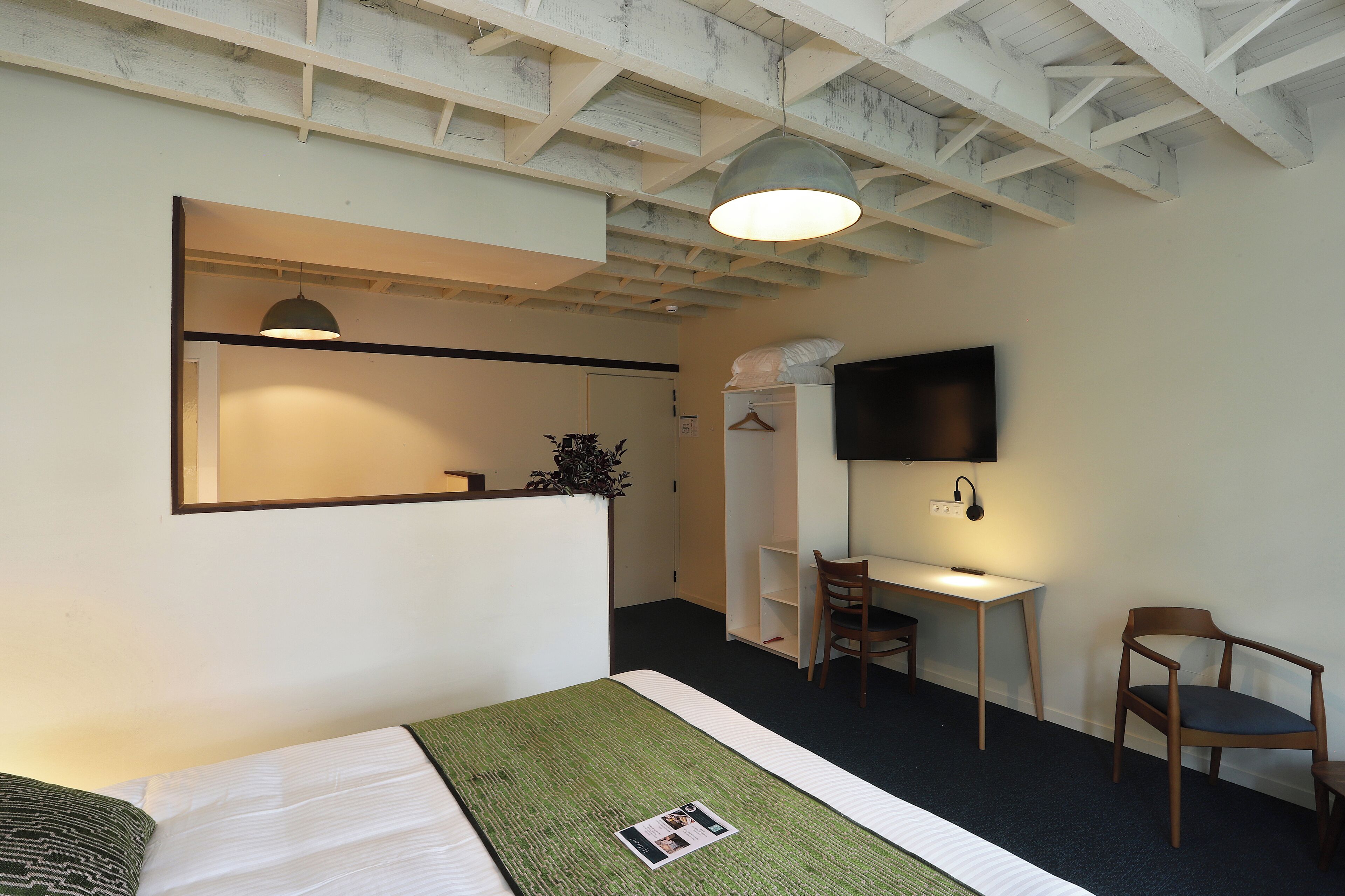 Ett hotellrum med en säng, ett skrivbord, en stol, en TV och ett tak med synliga takbjälkar.