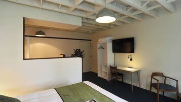 Ett hotellrum med en säng, ett skrivbord, en stol, en TV och ett tak med synliga takbjälkar.