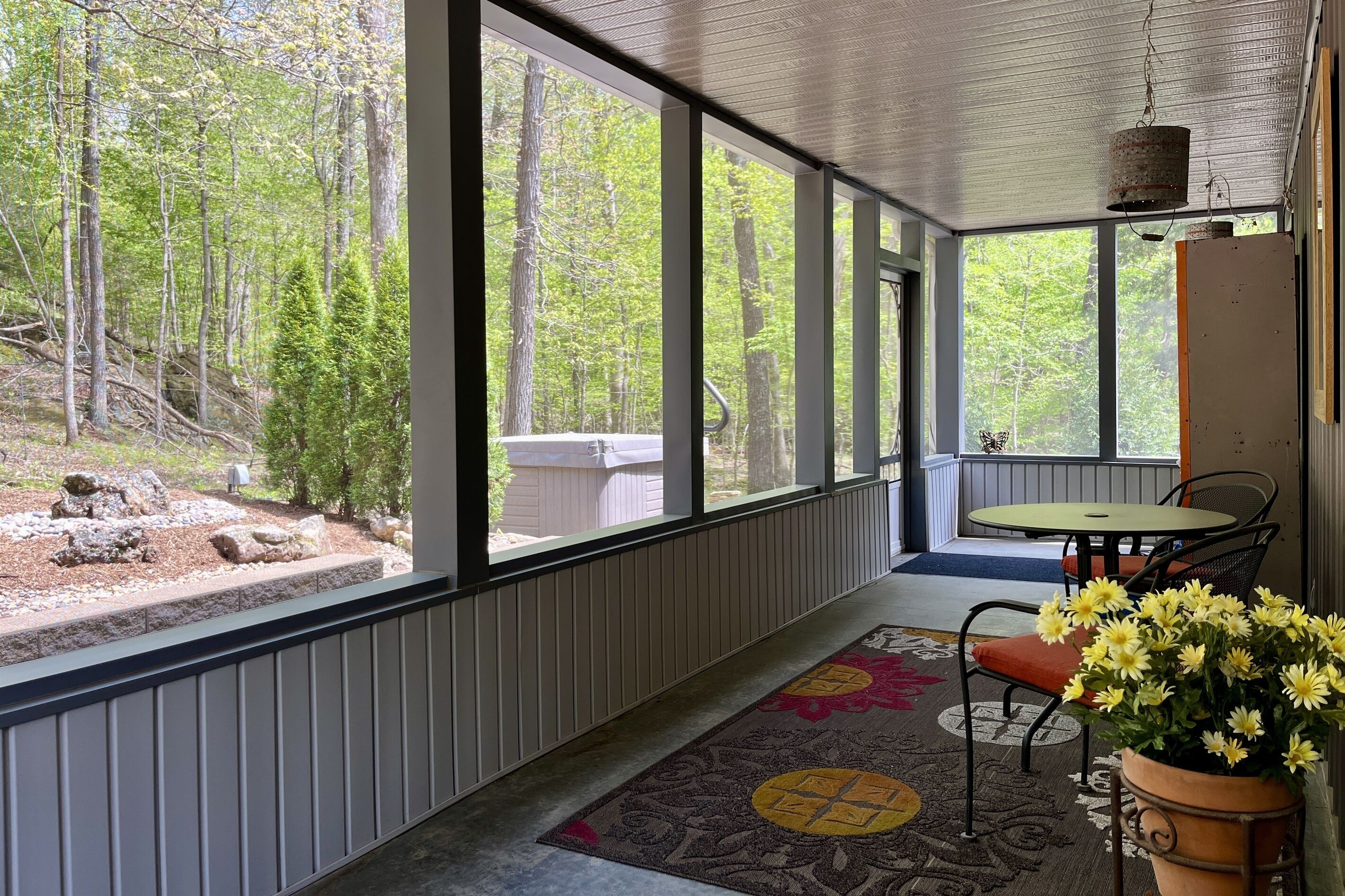 A covered porch with a view of a forest, a small table with chairs, and a potted plant with yellow flowers.