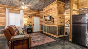 A cozy cabin-style room with a brown leather sofa, a small table, a TV mounted on a wooden wall, a fireplace, and a refrigerator.