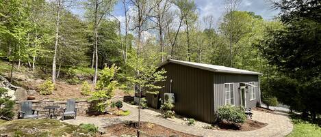 A small wooden cabin with a patio area, surrounded by trees and a stone pathway.