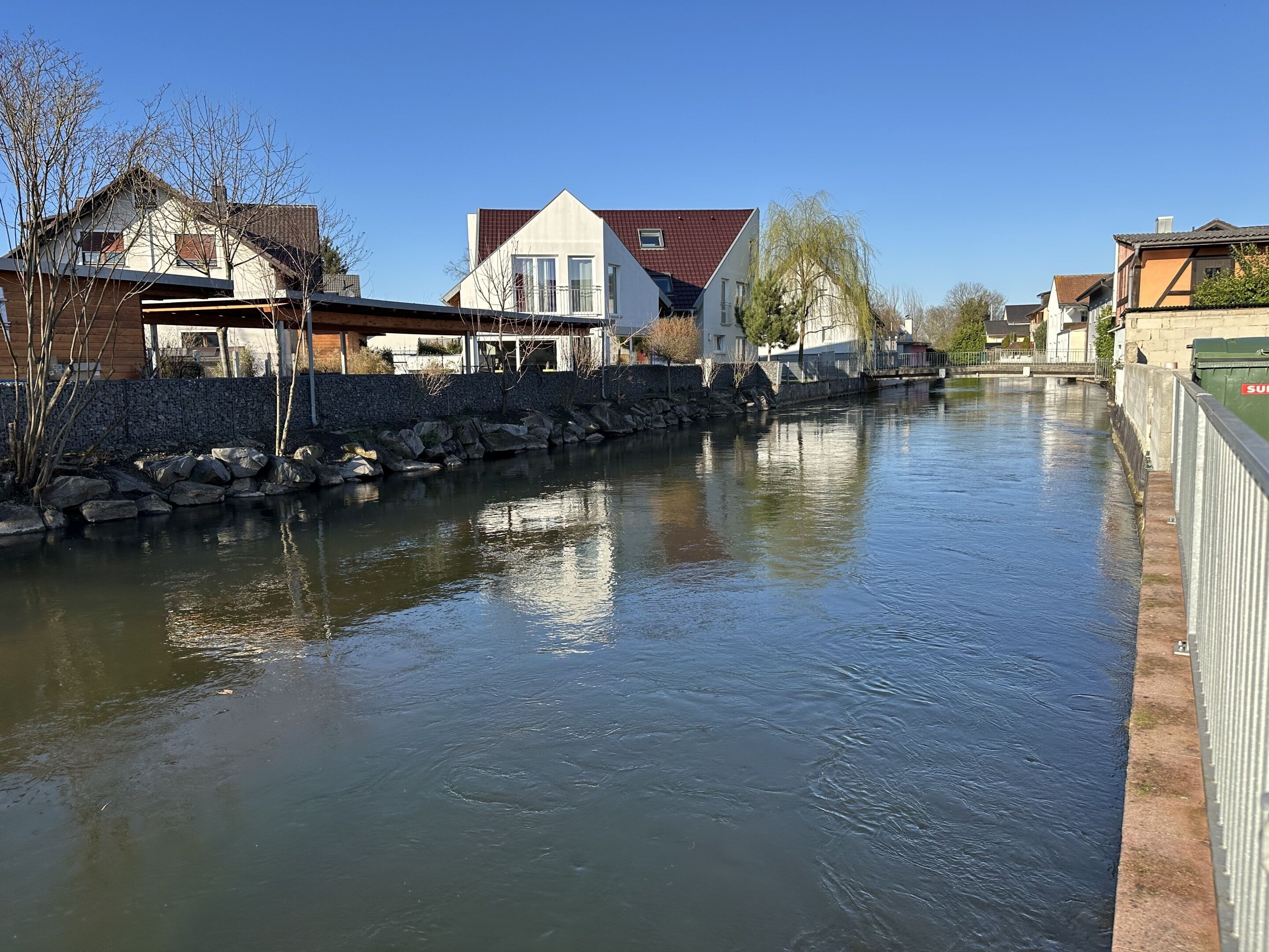 Ein ruhiger Fluss mit Häusern an einem Ufer und einer Steinmauer am anderen.