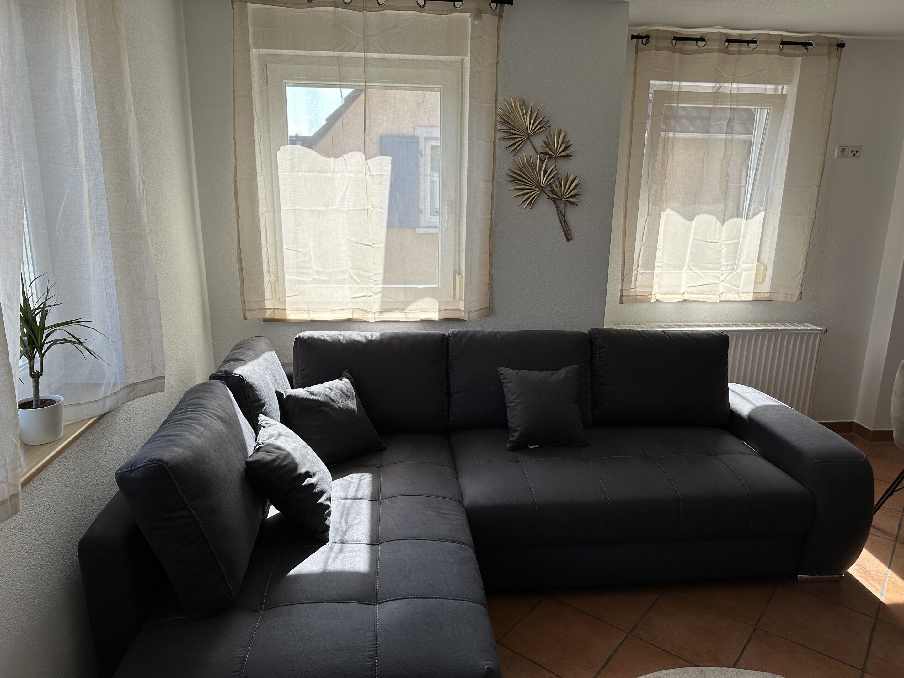 A living room with a sectional sofa, two windows with sheer curtains, a radiator, and a wall-mounted decorative piece.