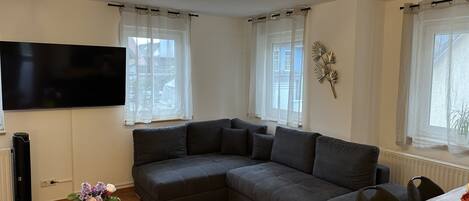 A modern living room with a sectional sofa, a flat-screen TV, and a wooden dining table.