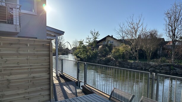 A wooden deck with a table and chairs overlooking a river, with buildings and trees in the background.