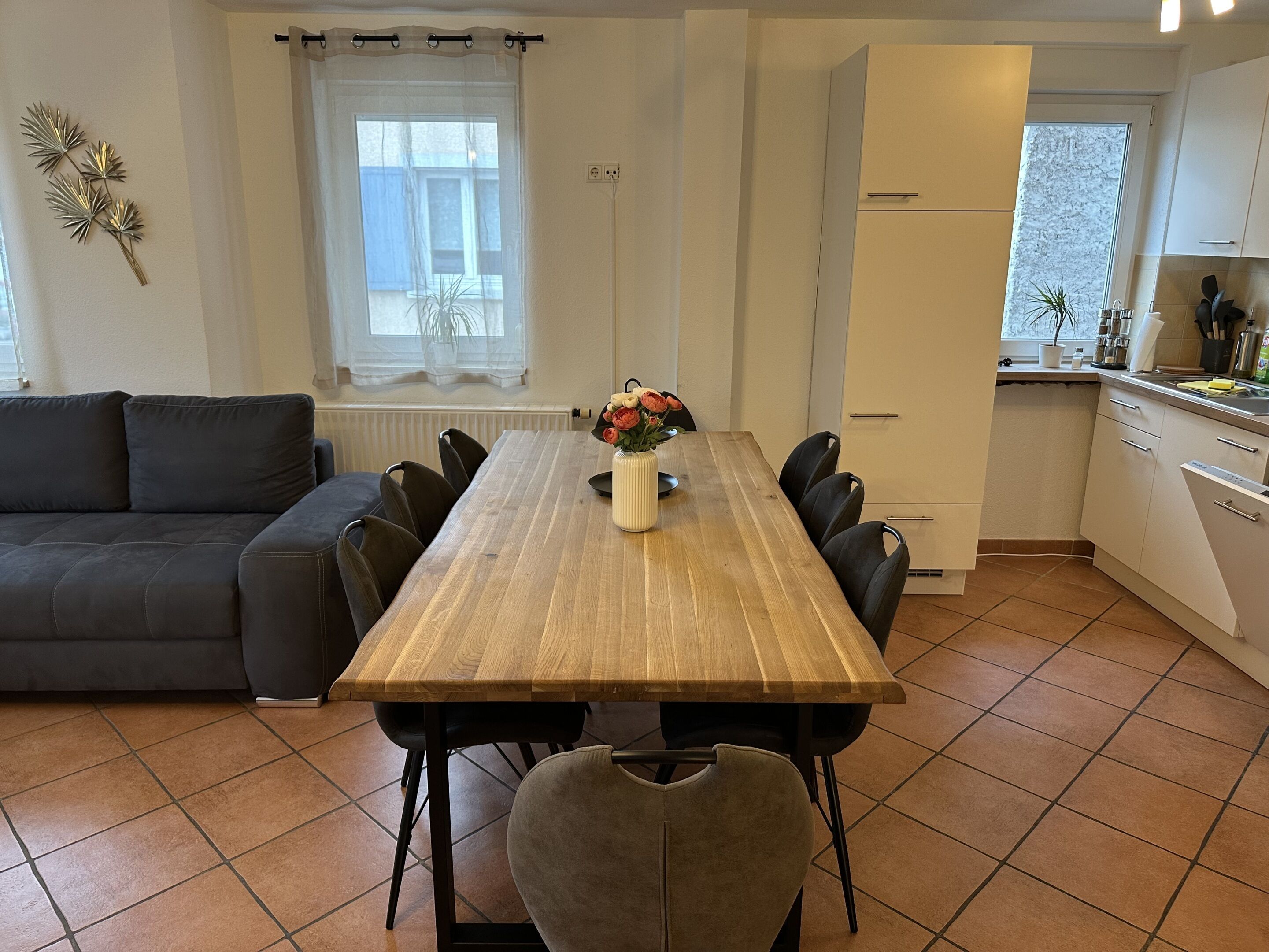 A dining area with a wooden table, chairs, a vase with flowers, a sofa, a kitchen with white cabinets, and a window with curtains.