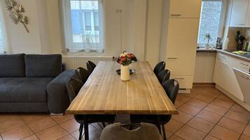 A dining area with a wooden table, chairs, a vase with flowers, a sofa, a kitchen with white cabinets, and a window with curtains.