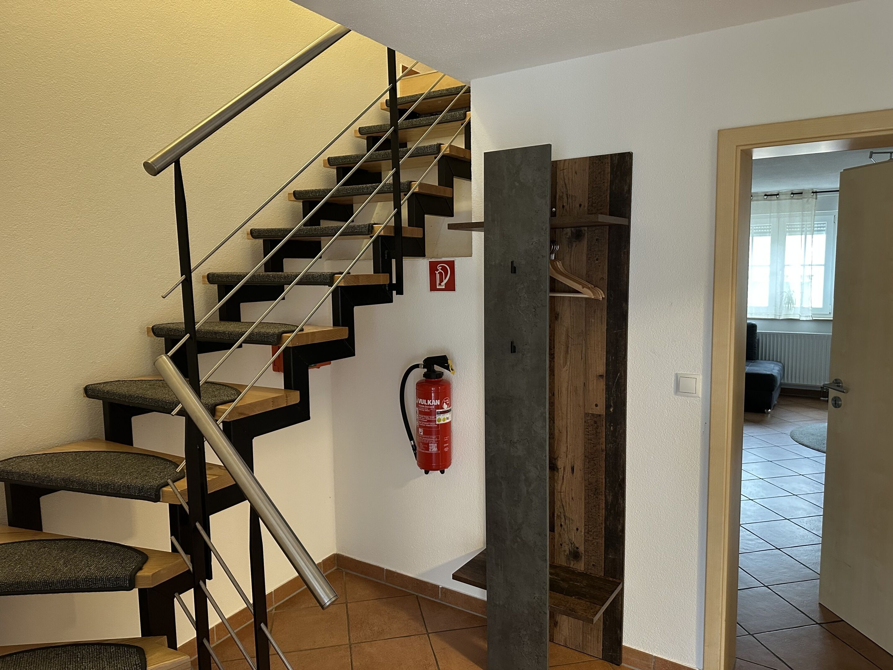 Una escalera con barandilla de metal y peldaños de madera, un extintor de incendios fijado a la pared y una puerta que da a otra habitación.