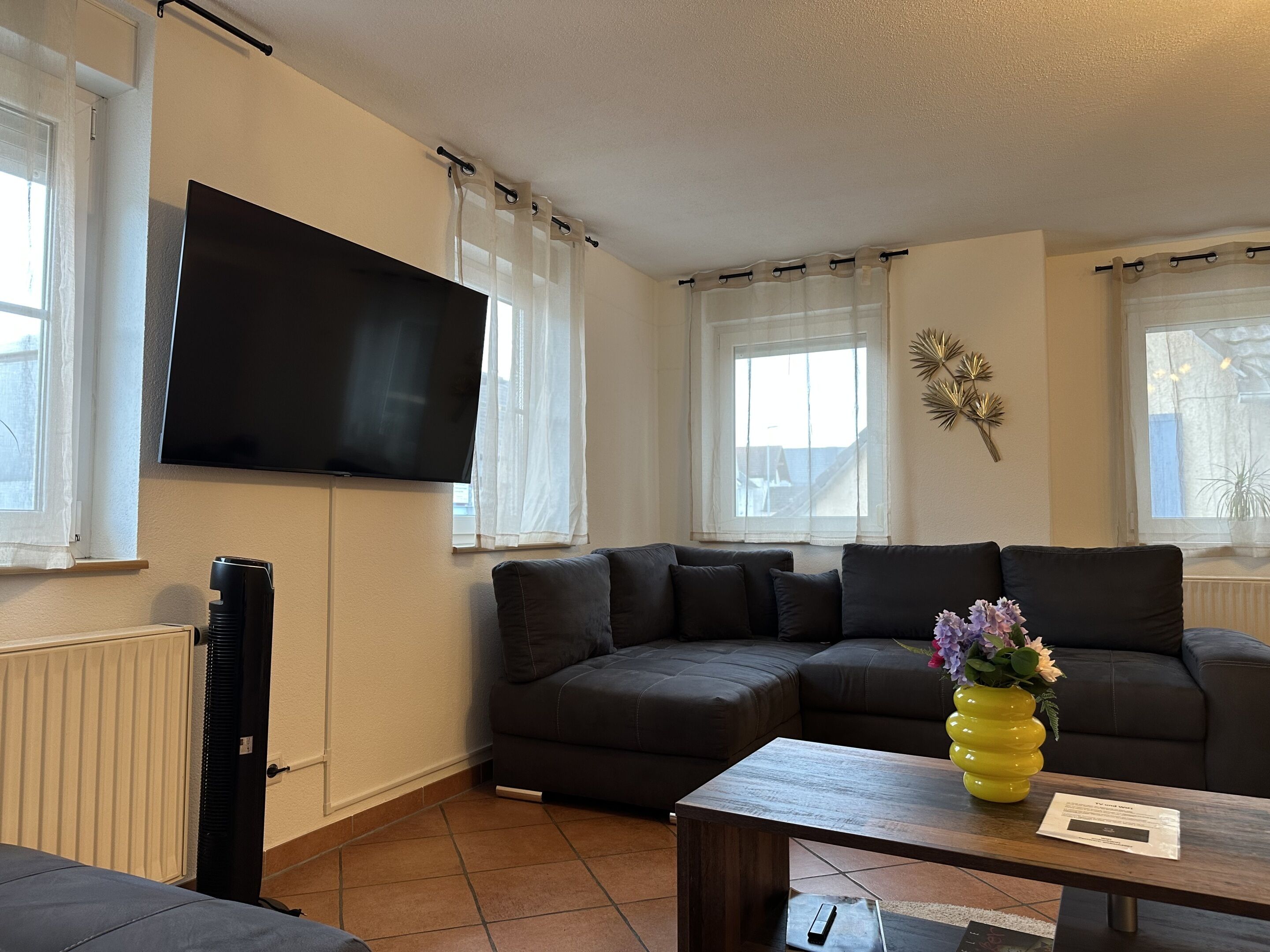 Ein Wohnzimmer mit Ecksofa, flachbildfernseher an der Wand, Couchtisch mit Vase und Heizkörper.