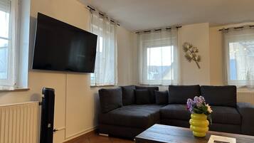 Ein Wohnzimmer mit Ecksofa, flachbildfernseher an der Wand, Couchtisch mit Vase und Heizkörper.